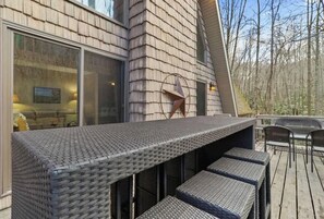 A modern outdoor bar area with wicker furniture and a large window overlooking a forested area. - A-Frame Of Mind: Mountain Escape (Beech Mountain)