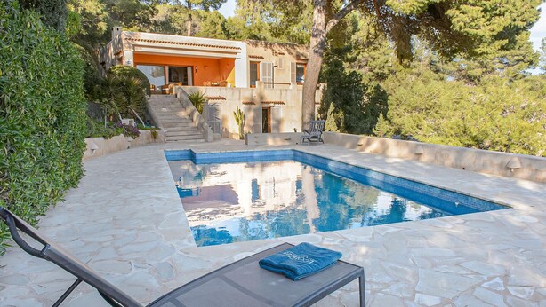 Piscina com deck de pedra, uma espreguiçadeira e um edifício com telhado cerâmico ao fundo.