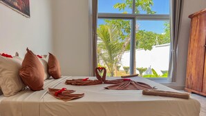 A bed with towels arranged in a heart shape, a wooden headboard, and a view of greenery outside. - EBENDE VILLA (Unguja South Region)