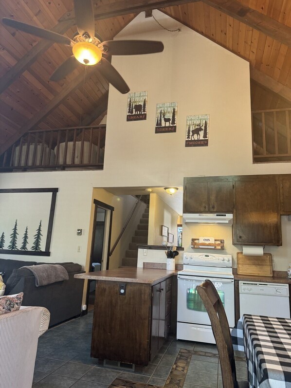 L’intérieur chaleureux d’un chalet avec un plafond en bois, un ventilateur de plafond, un coin cuisine équipé d’une cuisinière et de placards, une table à manger et un espace salon avec un canapé et un tableau représentant des arbres.