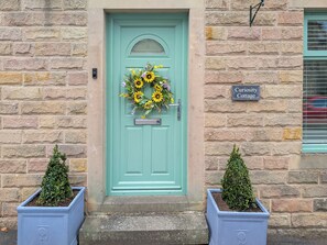 A turquoise door with a sunflowers wreath, a sign reading "Curiosity Cottage", and two potted plants on either side. - 3 bedroom accommodation in Eyam (Eyam)