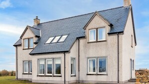 A two-story house with a steeply pitched roof, multiple windows, and a stone wall in the background. - 3 bedroom accommodation in South Boisdale (South Boisdale)