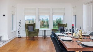 A dining area with a wooden table set for four, two green armchairs with patterned cushions, and a view of a green landscape through large windows. - 3 bedroom accommodation in South Boisdale (South Boisdale)