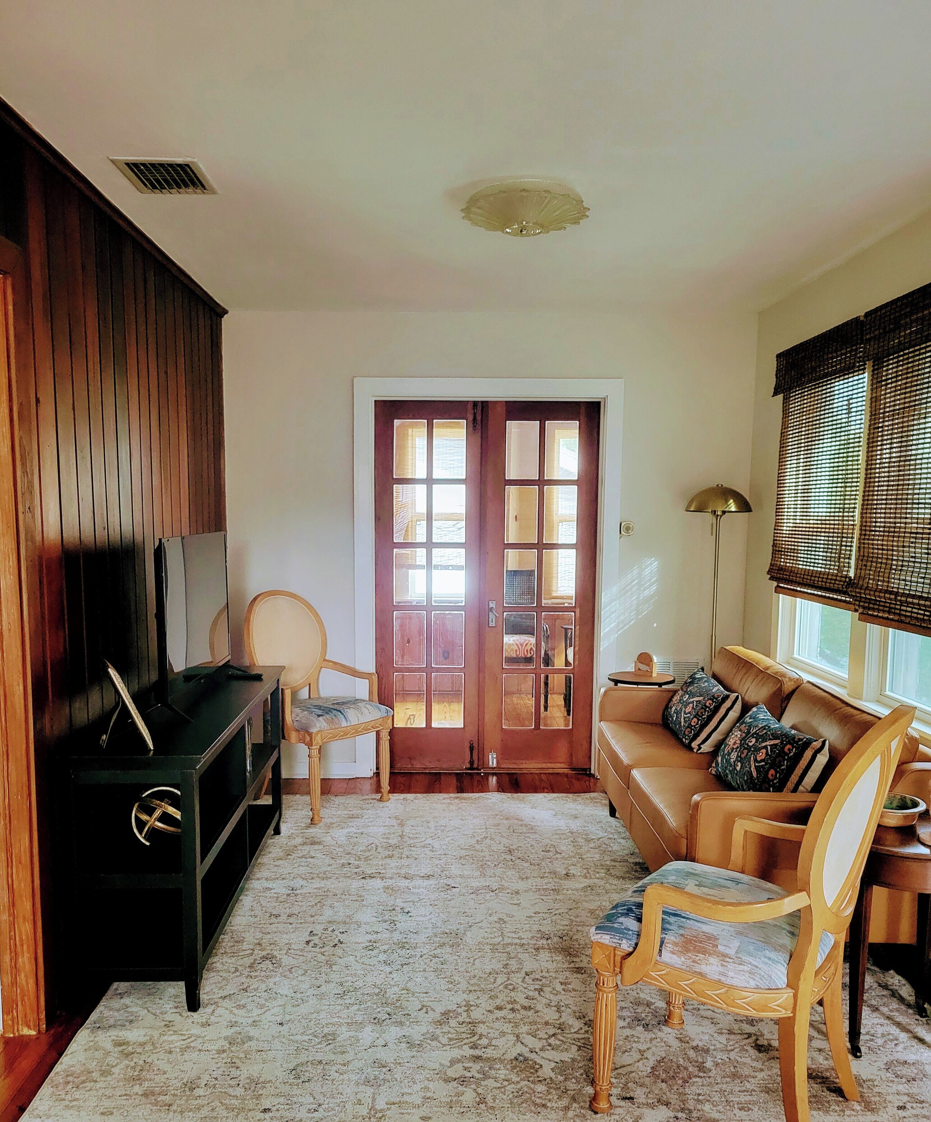 A living room with a wooden entertainment unit, a sofa, two chairs, a patterned rug, and a large window with blinds.