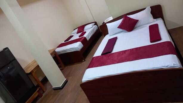 Habitación de hotel con dos camas individuales, cada una con una colcha roja y blanca y almohadas a juego. Hay una mesita de noche de madera con una lámpara y una pequeña mesa. El piso es de madera y las paredes son blancas.