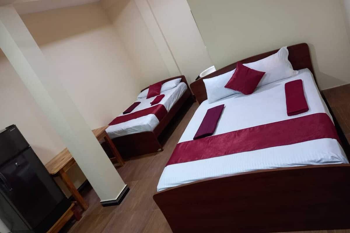 Habitación de hotel con dos camas individuales, cada una con una colcha roja y blanca y almohadas a juego. Hay una mesita de noche de madera con una lámpara y una pequeña mesa. El piso es de madera y las paredes son blancas.