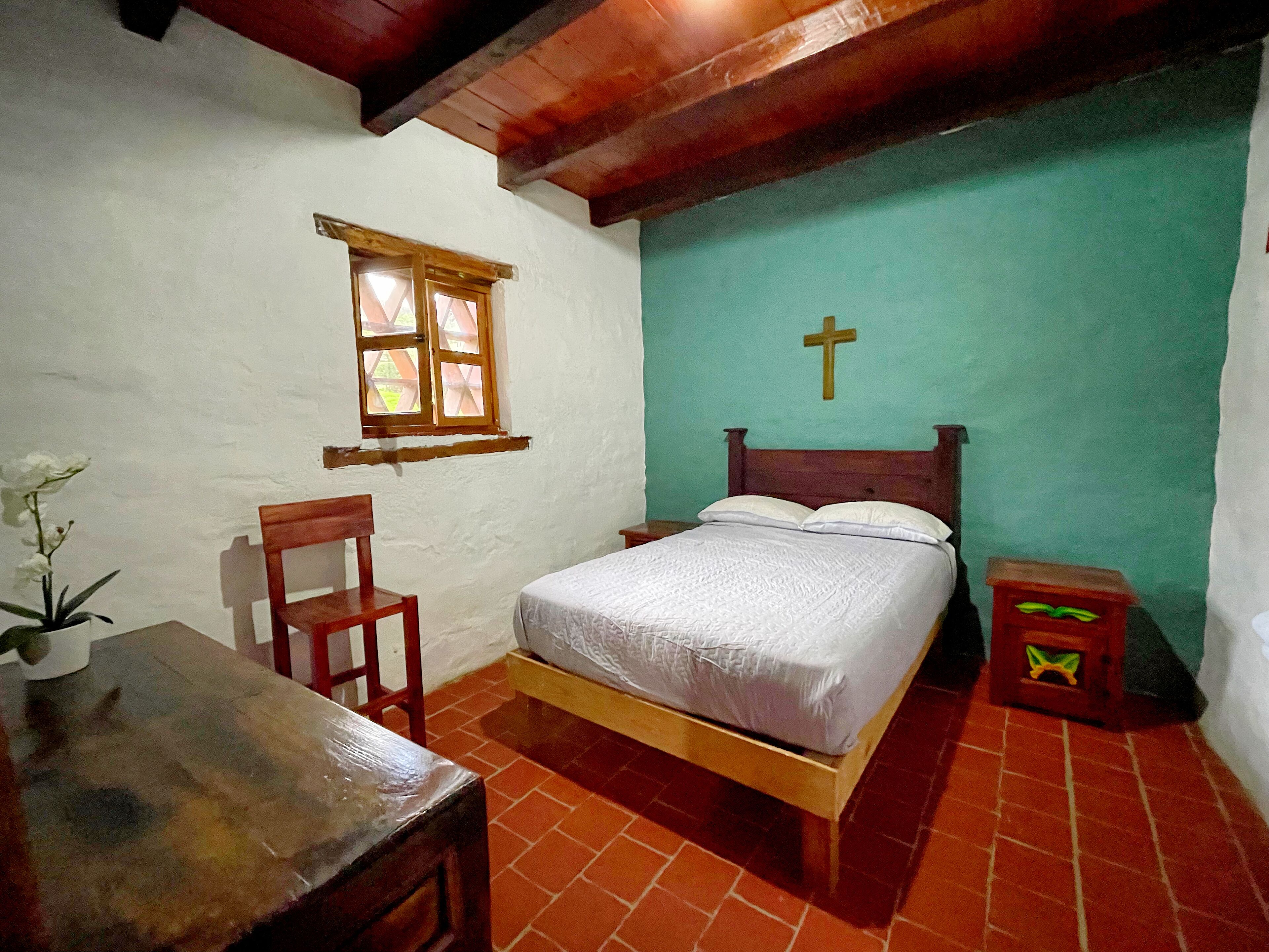 Un dormitorio pequeño y acogedor con una cama individual, una silla de madera, una mesita y una ventana. Hay una cruz en la pared.