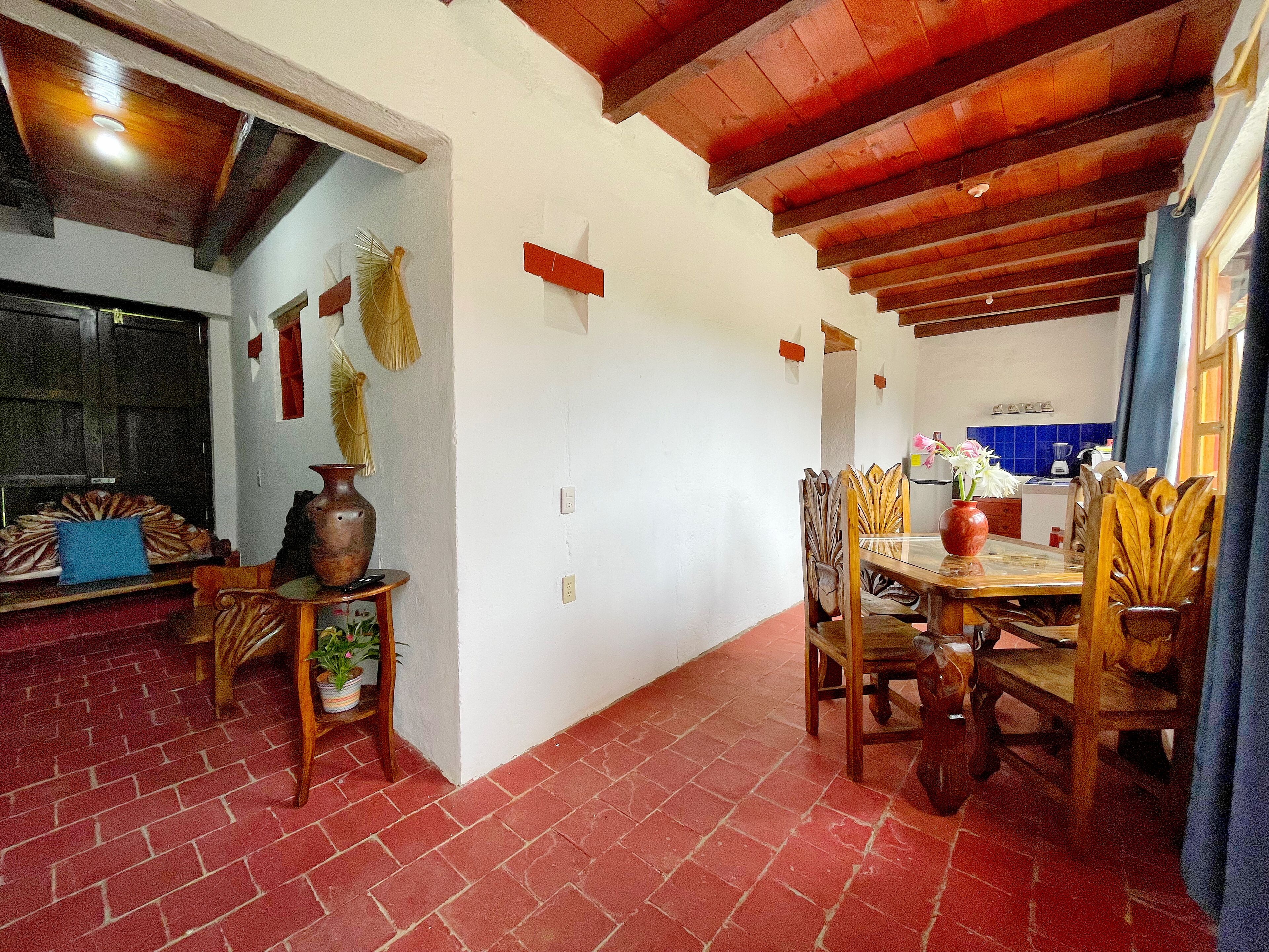 A dining area with a wooden table and chairs, a brick floor, and a kitchen in the background.