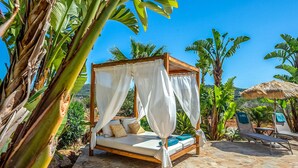 Un lit à baldaquin avec des rideaux blancs, une terrasse en pierre et un espace piscine avec des chaises longues.