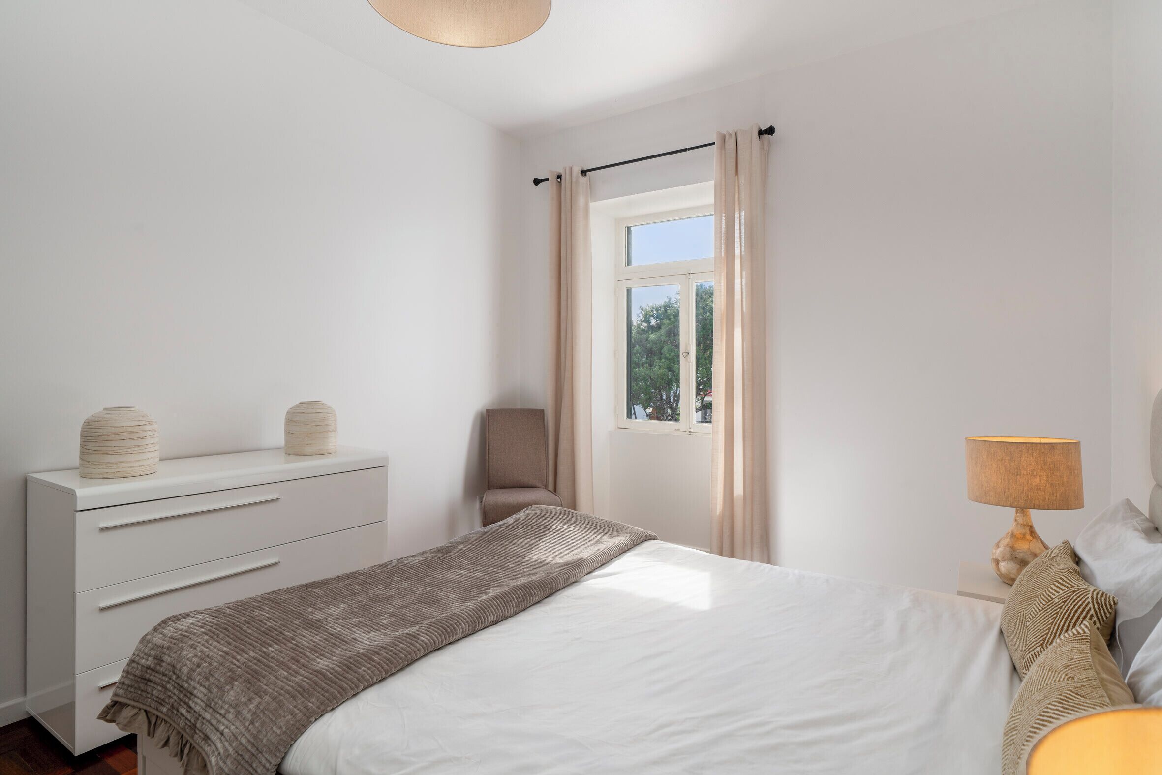 A bedroom with a bed, a dresser, a chair, a window with curtains, and a bedside lamp.