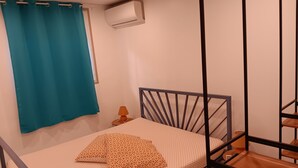 A single bed with a metal frame, a teal curtain, a small table with a lamp, and an air conditioning unit on the wall.