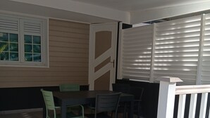 A covered patio area with a table and chairs, a white railing, and a window with blinds.
