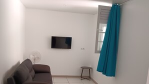 A living room with a dark brown sofa, a flat-screen TV mounted on the wall, a small round table, and a teal curtain.