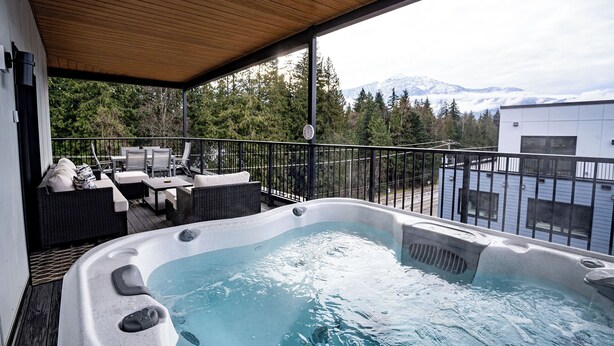 A hot tub on a covered deck with outdoor furniture, overlooking a mountainous landscape. - Alpine Retreat by Revelstoke Vacations (Revelstoke)