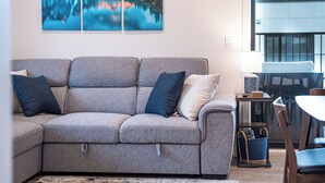 A living room with a grey sofa, two pillows, a small table with a lamp, and a view of mountains through a window. - Alpine Retreat by Revelstoke Vacations (Revelstoke)