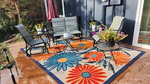 En udendørs terrasse med et blomsteret tæppe, stole og krukker med planter.
