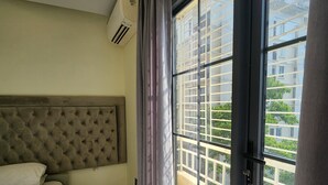 Habitación de hotel con cama con cabecera capitonada, una amplia puerta corrediza de vidrio a un balcón y un equipo de aire acondicionado en la pared.