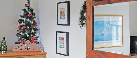 Un sapin de Noël décoré d’ornements et une petite maquette de maison sont posés sur un meuble en bois. Deux tableaux encadrés ornent le mur, et un miroir avec un cadre décoratif est également présent.