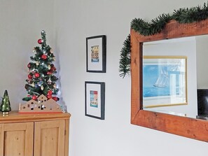 A Christmas tree with ornaments and a small house model on a wooden cabinet. There are two framed pictures on the wall and a mirror with a decorative frame. - We love this cosy cottage, located just a two minute walk from the beach. (Fareham)
