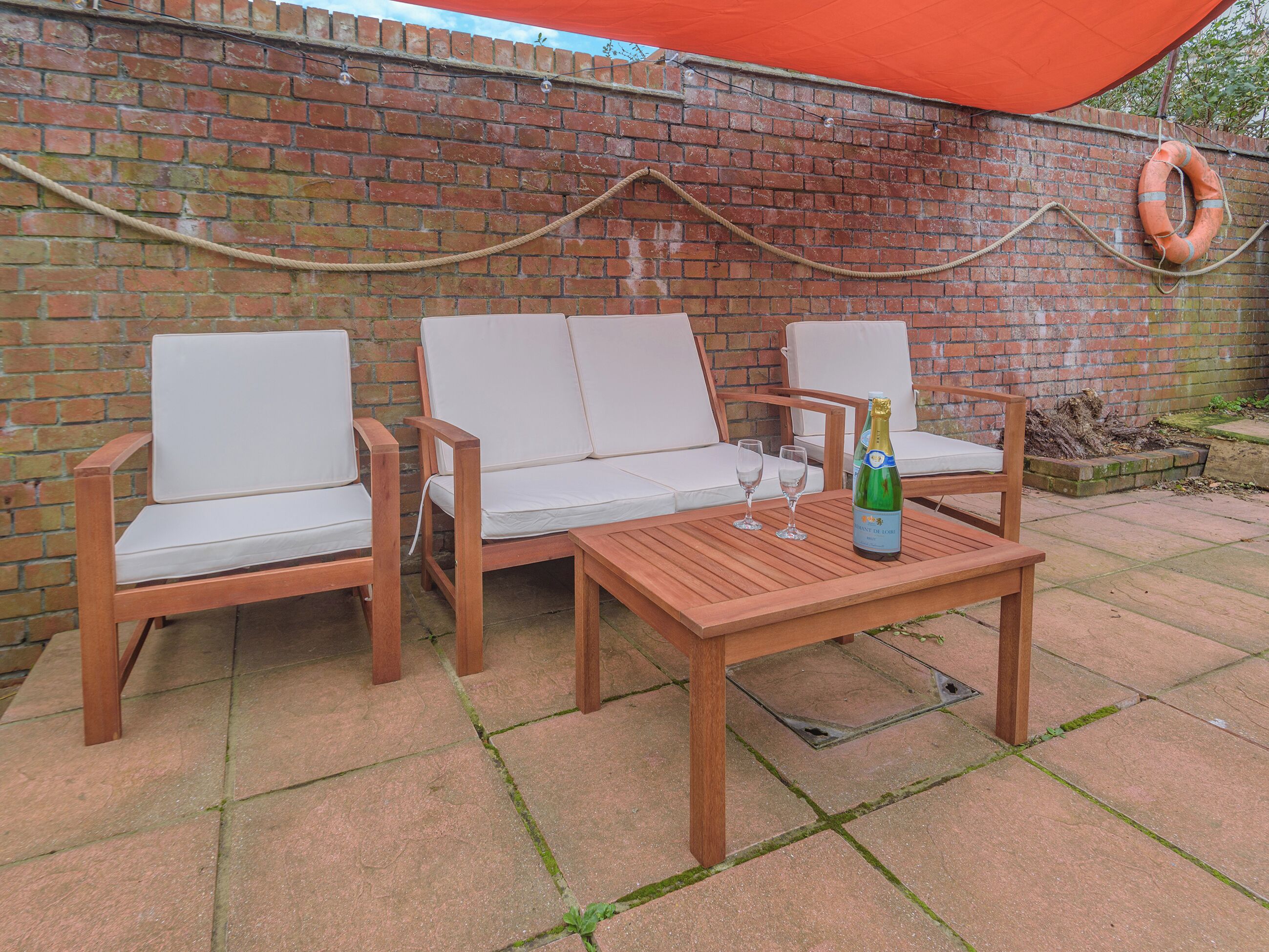 A patio with a brick wall, two white cushioned chairs, a wooden table, and a bottle of champagne.