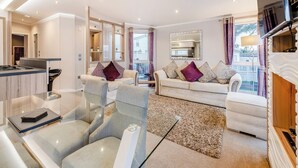 A modern living room with a sofa, chairs, a glass coffee table, a kitchen area, and a large window with curtains.