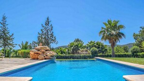 Una piscina rectangular con agua clara y azul, rodeada por un muro de piedra, palmeras y abundante vegetación.