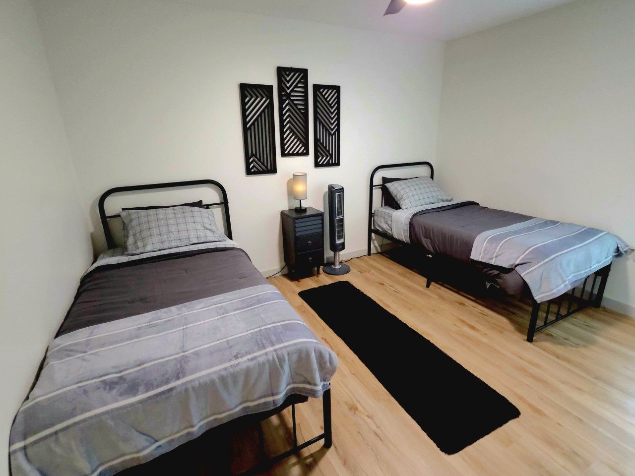Un dormitorio con dos camas, una mesita de noche con lámpara y una alfombra negra sobre piso de madera.