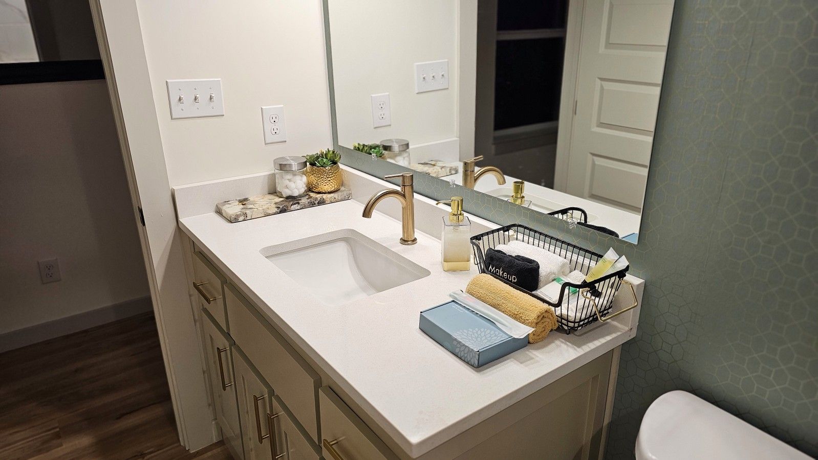 A bathroom with a white sink, gold faucet, and a mirror above it. There is a small plant on the counter and a basket with towels.