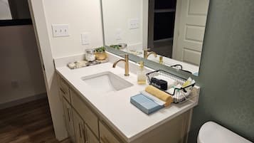 A bathroom with a white sink, gold faucet, and a mirror above it. There is a small plant on the counter and a basket with towels.