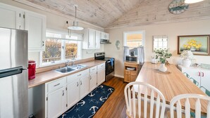 A kitchen with wooden cabinets, a stainless steel refrigerator, a sink, and a dining area with a wooden table and chairs. - Oceanside Cottage with Private Beach Access (Mattapoisett)