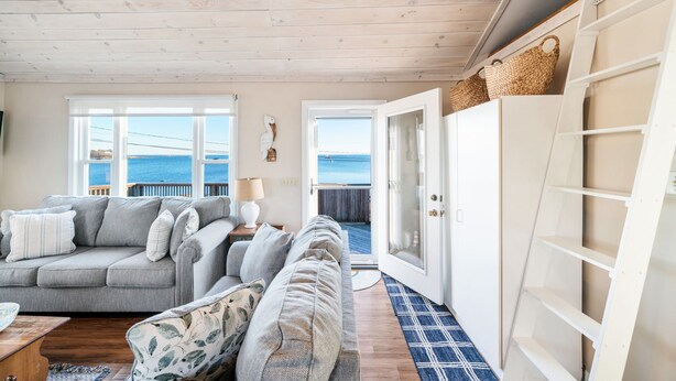 A cozy living room with a grey sofa, a patterned armchair, and a wooden ceiling. There is a large window with a view of the sea, and a white cabinet with baskets on top. - Oceanside Cottage with Private Beach Access (Mattapoisett)