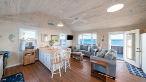 A spacious living area with a wooden ceiling, a dining table with chairs, a sofa, and a view of the sea through large windows. - Oceanside Cottage with Private Beach Access (Mattapoisett)