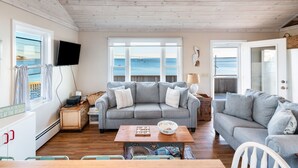 A cozy living room with a grey sofa, a wooden coffee table, and a television. Large windows offer a view of the sea. - Oceanside Cottage with Private Beach Access (Mattapoisett)