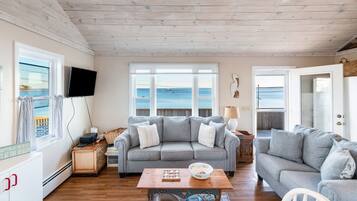 A cozy living room with a grey sofa, a wooden coffee table, and a television. Large windows offer a view of the sea.