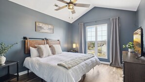 A bedroom with a bed, a ceiling fan, a window with curtains, a nightstand, and a plant. - NEW~9 min to Dollywood~Hot Tub~Arcade~4 Bedroom (Sevierville)