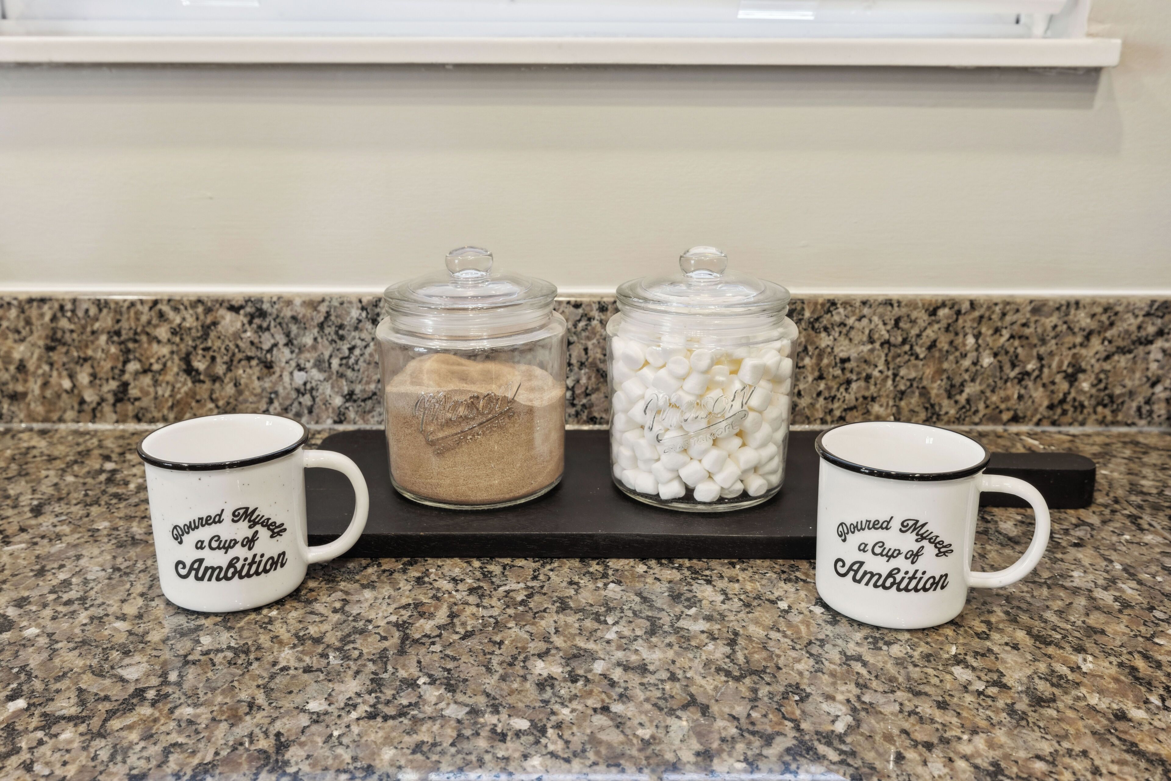 Two white mugs with black text on a granite countertop, each placed on a black tray.