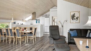 A modern kitchen with white cabinets, a dining area with wooden chairs and a table, and a grey armchair in a living area with a blue cushion. - Beautiful home in Vestervig with sauna (Vestervig)