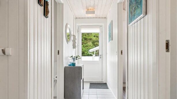 A hallway with white paneling, a door leading to a balcony, and a small table with blue items. - Beautiful home in Vestervig with sauna (Vestervig)