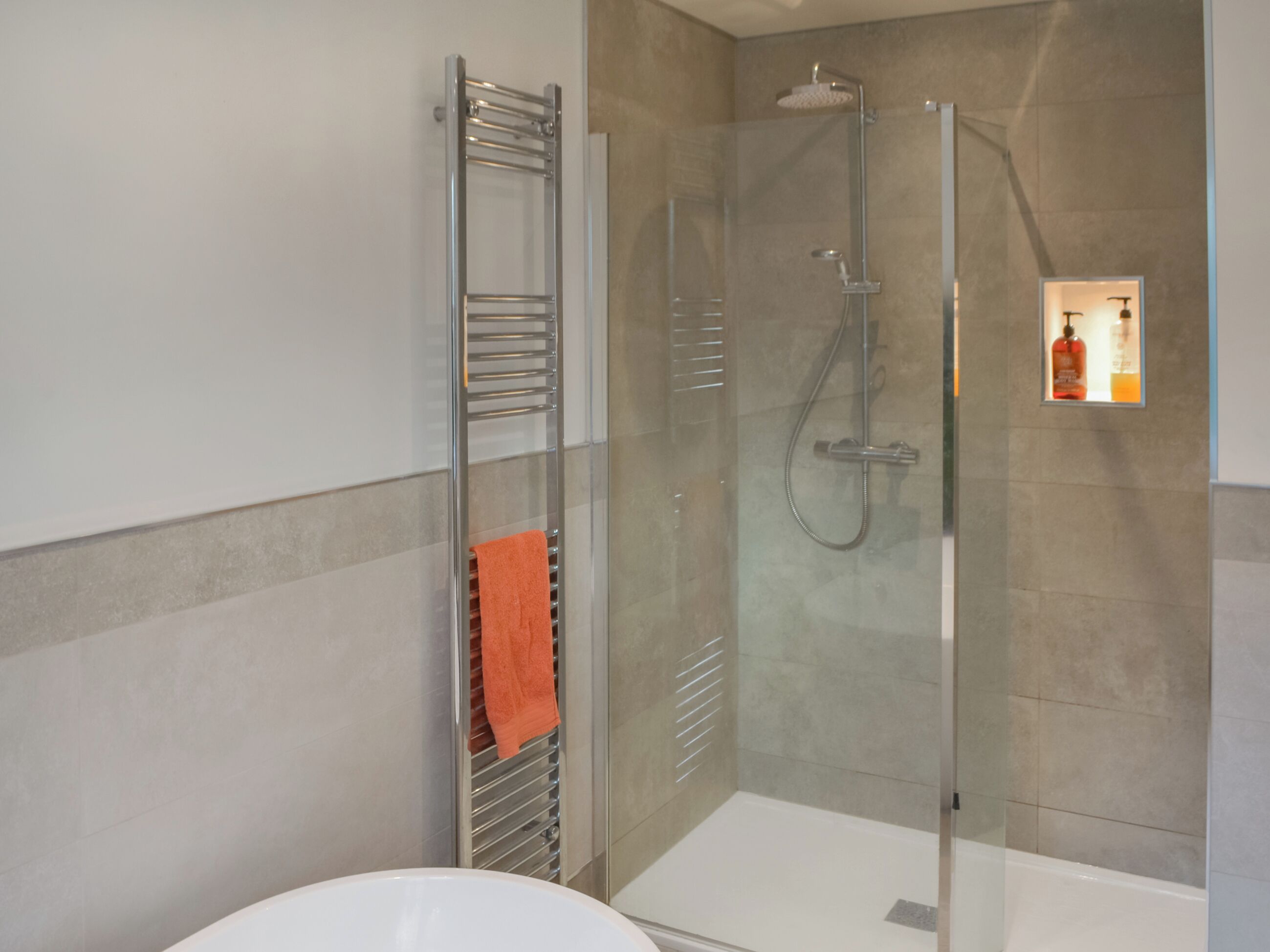 A modern bathroom with a glass-enclosed shower, a towel rack with an orange towel, and a bottle of shampoo on the wall.