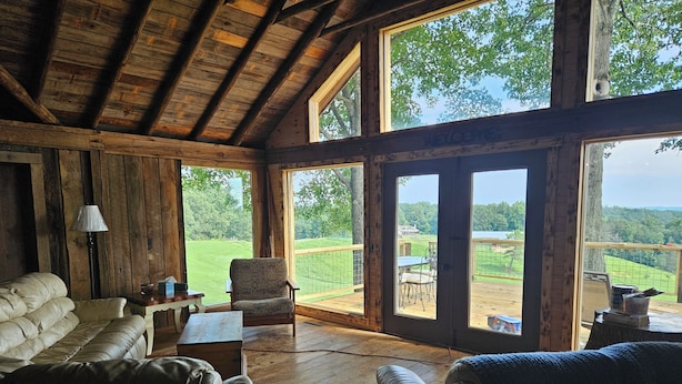 A cozy cabin interior with a wooden ceiling, large windows, a leather sofa, and a view of a green landscape. - Rustic Cabin on Homestead Farm (Dobson)