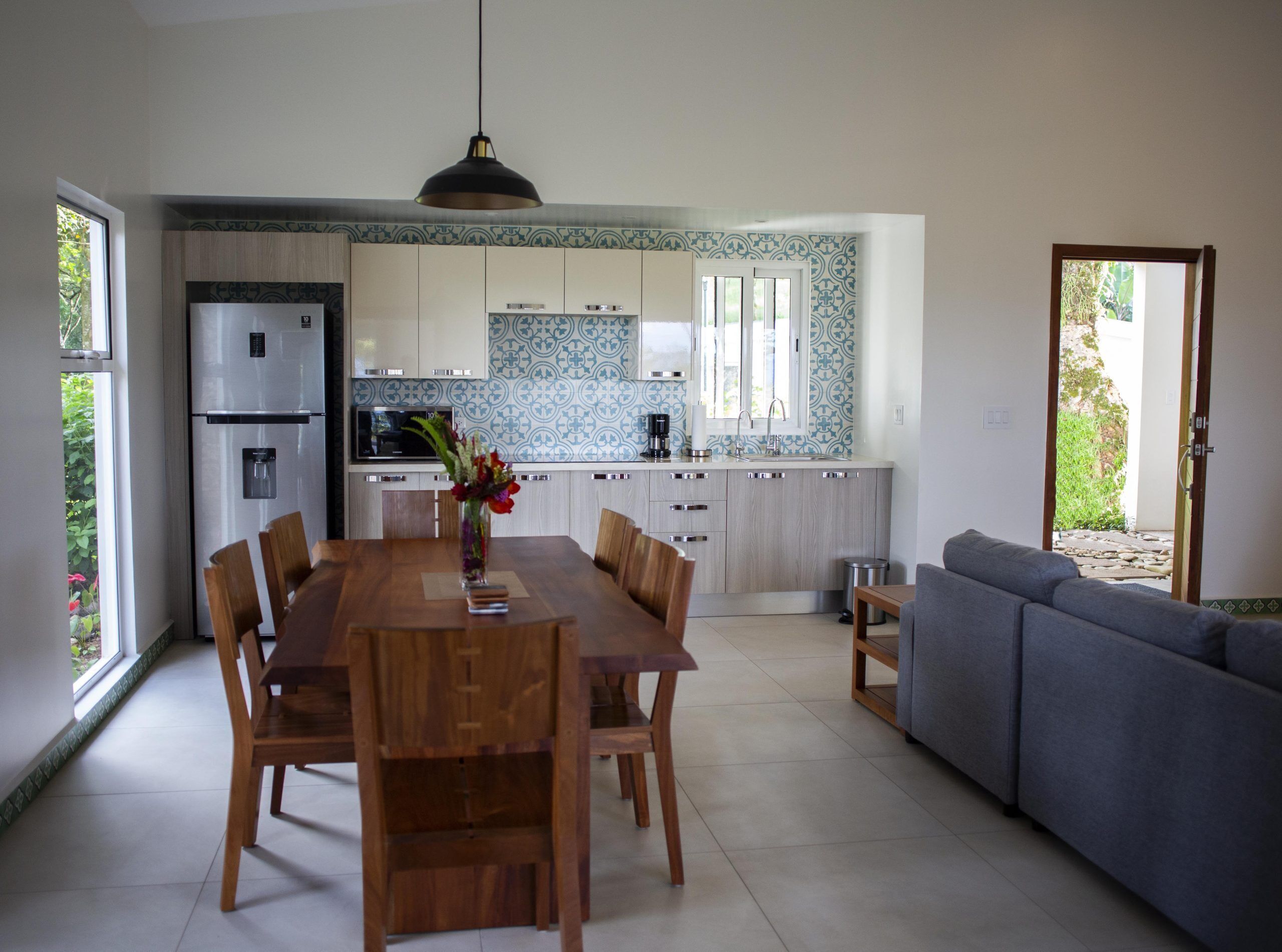 A modern kitchen with a central island, dining area with wooden furniture, and a grey sofa.
