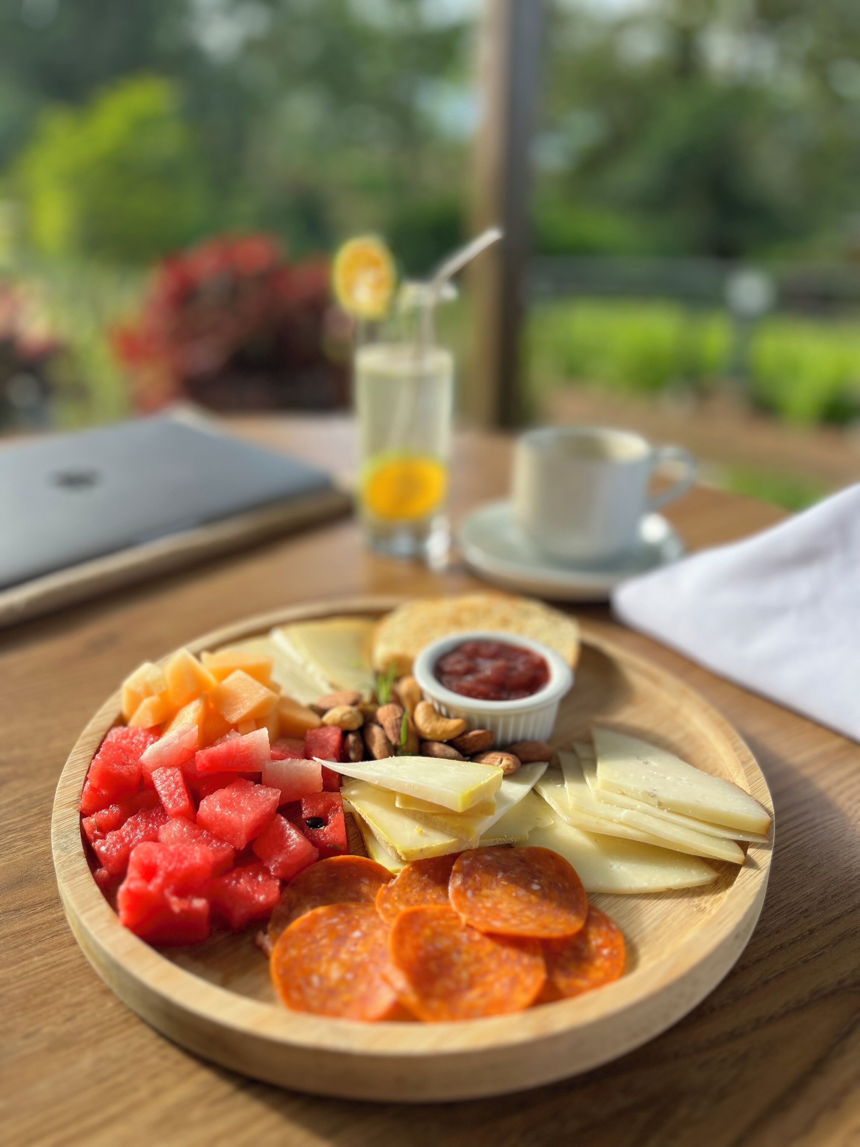 A wooden board with a variety of foods including watermelon, cheese, and nuts, and a cup of coffee on a table with a laptop and a glass of juice.
