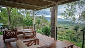 A wooden deck with outdoor furniture, including sofas and tables, overlooking a lush green landscape with trees and mountains.