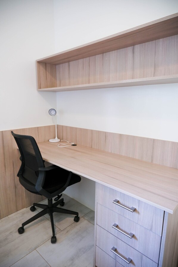 A corner office with a wooden desk, black office chair, and a white desk lamp. There are three drawers with silver handles.