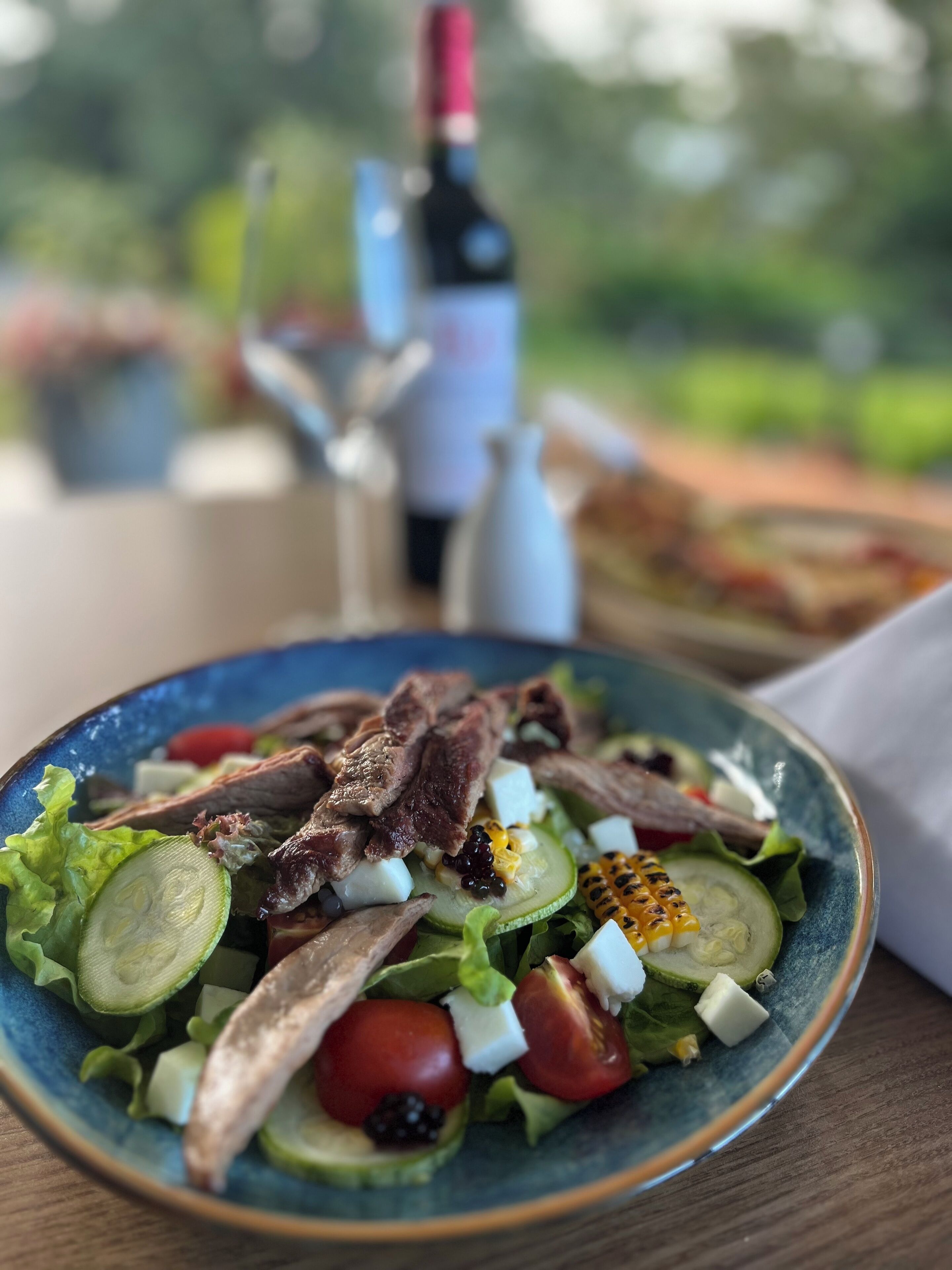 A bowl of salad with various ingredients, a bottle of wine, and a glass on a table.