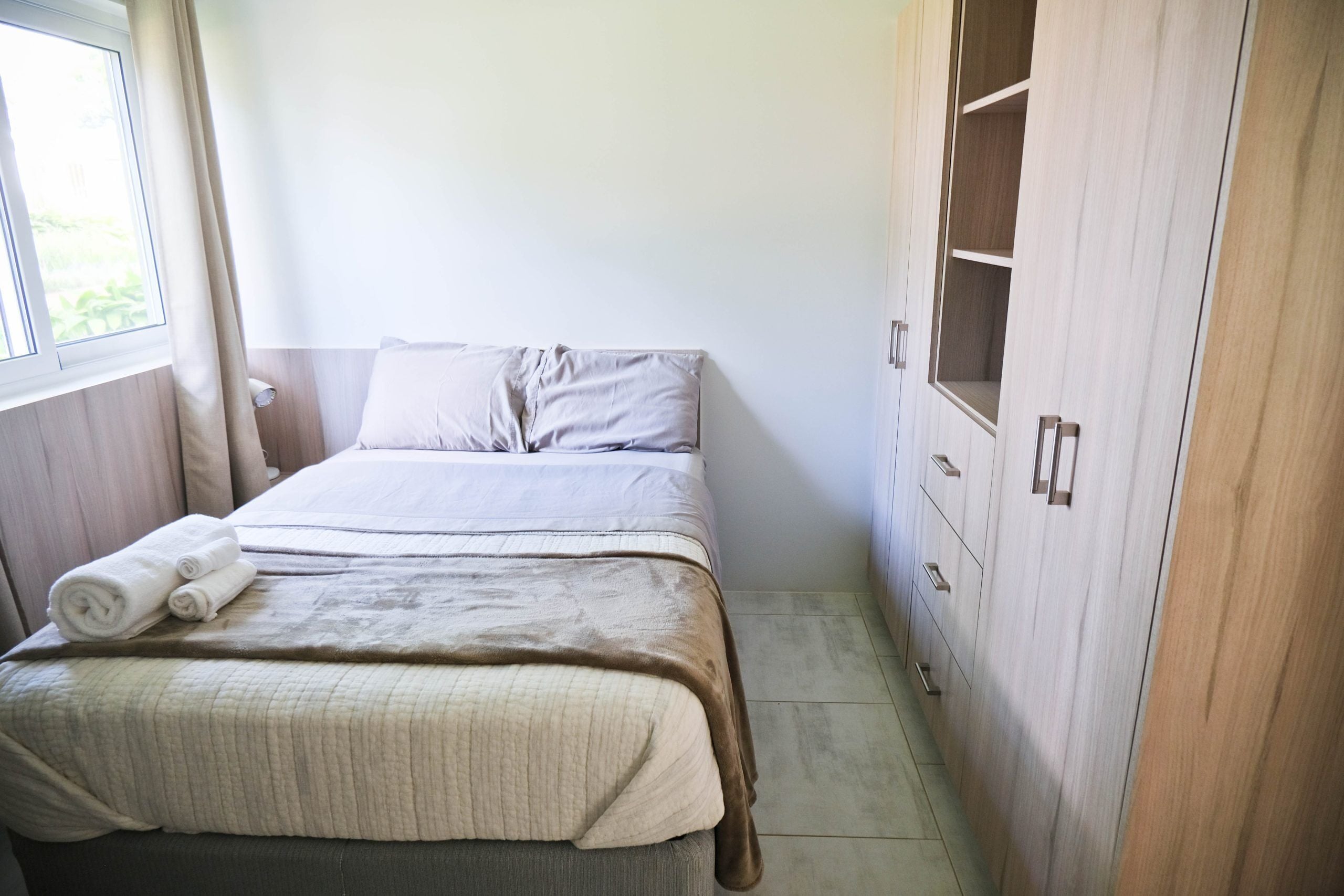 Una cama bien hecha con una manta color beige y almohadas, un juego de toallas y un armario de madera con varios compartimentos.