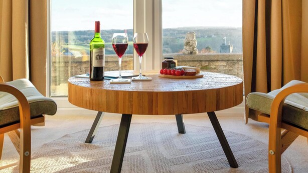 A round wooden table with a bottle of wine, two wine glasses, and a plate of food.