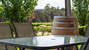 An outdoor patio with a glass table and metal chairs, surrounded by greenery and a wooden barrel.