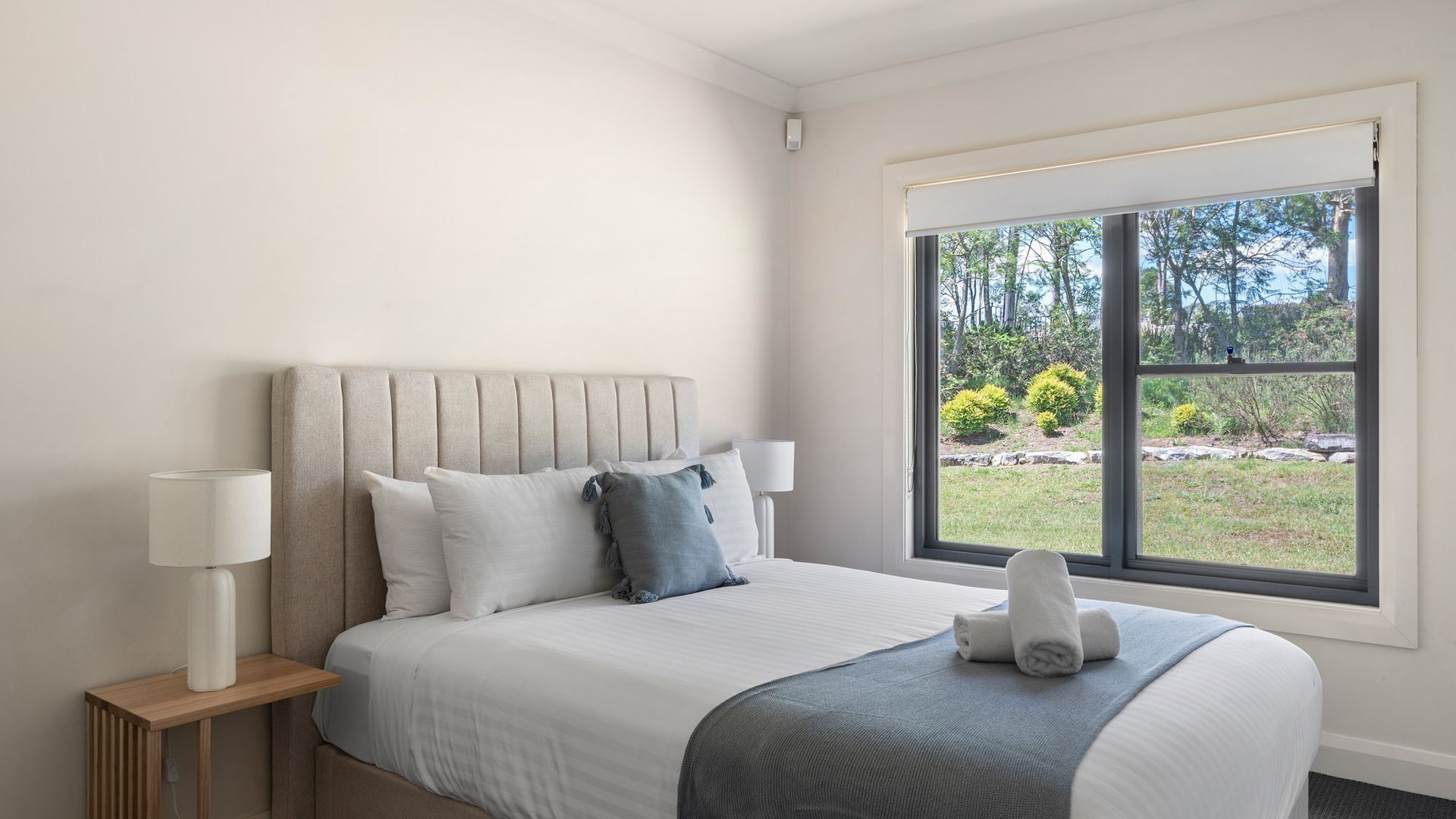 A hotel room with a large window, a bed with white bedding, two bedside lamps, and a view of a garden.
