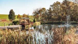 Une terrasse en bois rustique surplombe un étang agrémenté d’une fontaine, le tout étant entouré de hautes herbes et d’arbres.